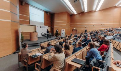 Duża sala wykładowa wypełniona ludźmi siedzącymi i zwróconymi w stronę sceny, na której prelegent stoi w pobliżu podium i ekranu projekcyjnego na Kongresie Strategicznym. Publiczność wydaje się być uważna. Sala jest wyłożona drewnianą boazerią, ma wielopoziomowe siedzenia i jasne oświetlenie sufitowe.