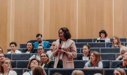 Kobieta w jasnoróżowej marynarce stoi i trzyma mikrofon, przemawiając na Kongresie Strategicznym do siedzącej publiczności w sali wykładowej. Publiczność, złożona z mężczyzn i kobiet, słucha uważnie wśród drewnianych paneli i wielopoziomowych szarych siedzeń.