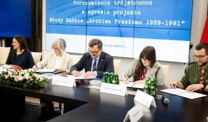 Pięć osób siedzi przy stole konferencyjnym i podpisuje dokumenty. Na dużym ekranie za nimi wyświetlany jest polski tekst o projekcie 