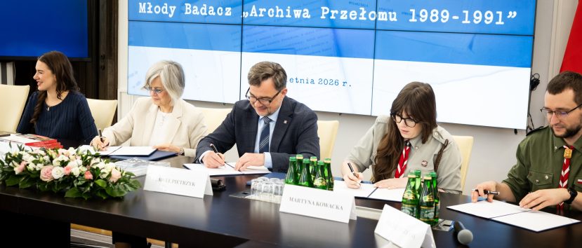 Pięć osób siedzi przy stole konferencyjnym i podpisuje dokumenty. Na dużym ekranie za nimi wyświetlany jest polski tekst o projekcie 