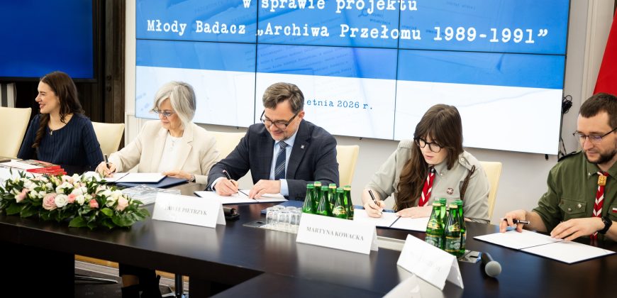 Pięć osób siedzi przy stole konferencyjnym i podpisuje dokumenty. Na dużym ekranie za nimi wyświetlany jest polski tekst o projekcie 