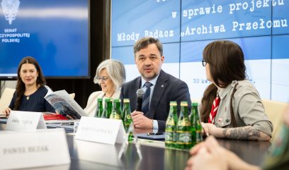 Pięć osób siedzi przy stole konferencyjnym z butelkami wody i dokumentami. Mężczyzna w garniturze mówi do mikrofonu. Duże ekrany za nimi wyświetlają polski tekst i logo. Atmosfera jest formalna i profesjonalna, co wskazuje na oficjalną dyskusję lub spotkanie.
