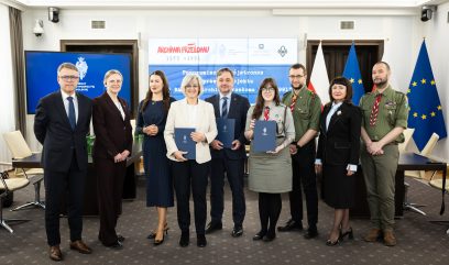 Dziewięć osób stoi w środku w mundurach formalnych i harcerskich, trzymając niebieskie teczki. Za nimi znajdują się flagi Polski i UE oraz ekran wyświetlający polski tekst. Wszyscy są uśmiechnięci, ustawieni obok siebie jak podczas konferencji lub ceremonii wręczenia nagród.
