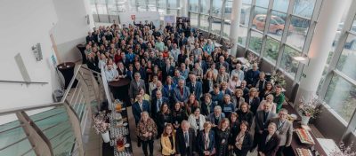 Duża grupa ludzi, ubranych głównie w stroje biznesowe, stoi blisko siebie w jasnym, nowoczesnym atrium z wysokimi oknami podczas Kongresu Strategicznego. Przestrzeń zdobią stoły z przekąskami i kwiatami. Zdjęcie zostało zrobione z góry na klatce schodowej.