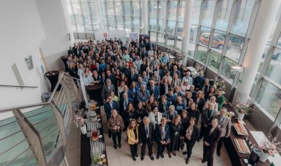 Duża grupa ludzi, ubranych głównie w stroje biznesowe, stoi blisko siebie w jasnym, nowoczesnym atrium z wysokimi oknami podczas Kongresu Strategicznego. Przestrzeń zdobią stoły z przekąskami i kwiatami. Zdjęcie zostało zrobione z góry na klatce schodowej.