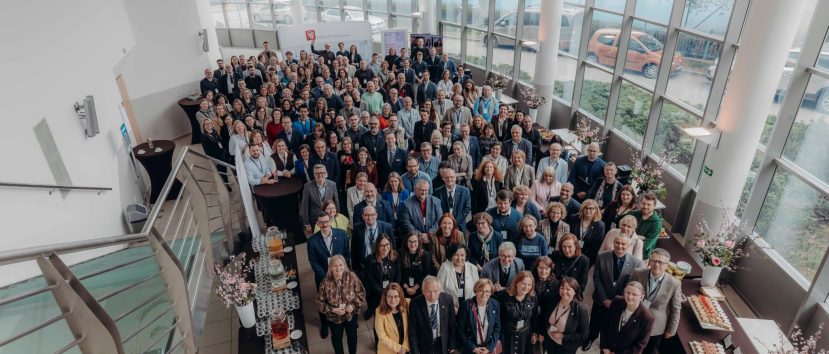 Duża grupa ludzi, ubranych głównie w stroje biznesowe, stoi blisko siebie w jasnym, nowoczesnym atrium z wysokimi oknami podczas Kongresu Strategicznego. Przestrzeń zdobią stoły z przekąskami i kwiatami. Zdjęcie zostało zrobione z góry na klatce schodowej.