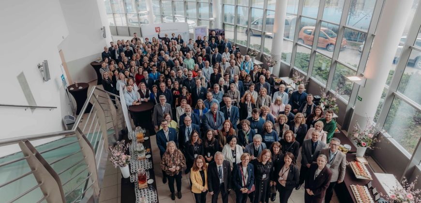 Duża grupa ludzi, ubranych głównie w stroje biznesowe, stoi blisko siebie w jasnym, nowoczesnym atrium z wysokimi oknami podczas Kongresu Strategicznego. Przestrzeń zdobią stoły z przekąskami i kwiatami. Zdjęcie zostało zrobione z góry na klatce schodowej.