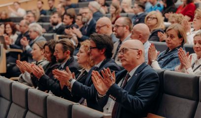 Duża grupa ludzi siedzi w rzędach w audytorium, ubrana w strój biznesowy lub formalny. Skupiają się do przodu i klaszczą, sugerując, że oglądają prezentację Kongresu Strategicznego. Publiczność wydaje się zaangażowana i uważna.