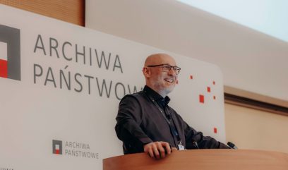 Uśmiechnięty, łysy mężczyzna w okularach i z brodą stoi na drewnianym podium, prowadząc prezentację na Kongresie Strategicznym. Za nim znajduje się biały baner z napisem ARCHIWA PAŃSTWOWE i czerwono-szarym logo, sugerujący miejsce konferencji lub seminarium.