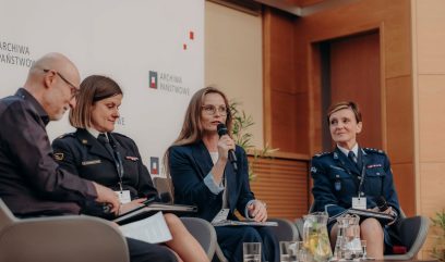 Podczas panelu dyskusyjnego Kongresu Strategicznego na scenie siedzą cztery osoby: dwie kobiety w policyjnych mundurach, kobieta w niebieskim garniturze trzymająca mikrofon oraz mężczyzna w okularach i z teczkami. Na stole stoją szklanki z wodą, a w tle widnieje napis 