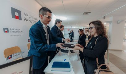 Mężczyzna w niebieskim garniturze wręcza dokument uśmiechniętej kobiecie przy stanowisku rejestracji podczas Kongresu Strategicznego. Kilka osób stoi w jasnym, nowoczesnym lobby z napisem 