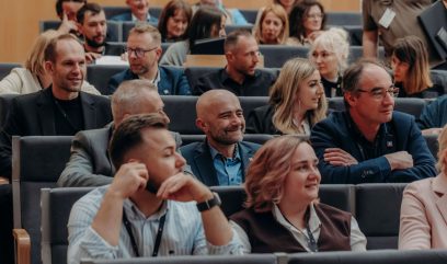Grupa mężczyzn i kobiet siedzi blisko siebie w rzędach siedzeń w audytorium, twarzami do przodu. Większość z nich uśmiecha się lub rozmawia, ubrana w strój biznesowy lub elegancki, co sugeruje, że uczestniczą w konferencji Kongres Strategiczny.