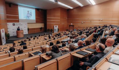 Duża sala wykładowa z drewnianymi ścianami jest wypełniona uczestnikami Kongresu Strategicznego, którzy stoją przed prelegentem na podium. Na ekranie wyświetlana jest prezentacja przedstawiająca zdjęcie dziecka, podczas gdy niektórzy uczestnicy robią notatki na swoich laptopach.