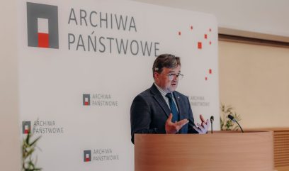 Mężczyzna w garniturze i krawacie przemawia na podium z mikrofonami. Za nim widnieje duży napis 