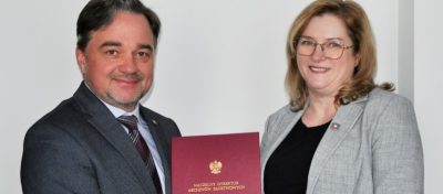 Dwie osoby w szarych marynarkach stoją obok siebie, uśmiechając się. Mężczyzna po lewej stronie trzyma czerwoną teczkę ze złotym polskim orłem i tekstem. Kobieta po prawej ma blond włosy i okulary. Oboje stoją przed białą ścianą.