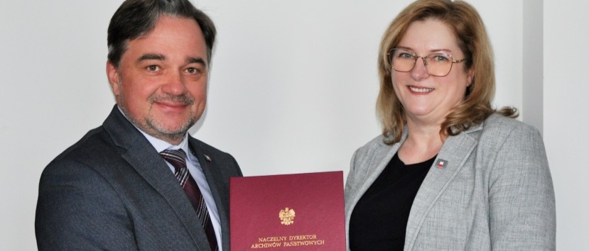 Dwie osoby w szarych marynarkach stoją obok siebie, uśmiechając się. Mężczyzna po lewej stronie trzyma czerwoną teczkę ze złotym polskim orłem i tekstem. Kobieta po prawej ma blond włosy i okulary. Oboje stoją przed białą ścianą.