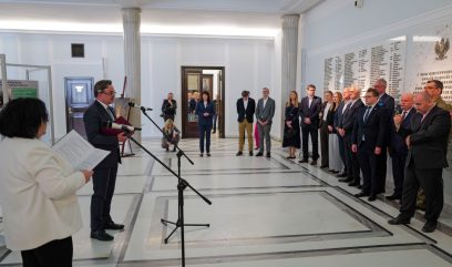 Mężczyzna przemawia do mikrofonu w jasnej, formalnej sali, podczas gdy kobieta stoi obok, trzymając dokumenty. Grupa ludzi w garniturach i formalnych strojach stoi uważnie po prawej stronie. W tle widoczne są kolumny, tablice i wysoki sufit.