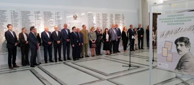 Grupa ludzi w oficjalnych strojach stoi w szeregu wewnątrz jasnej sali z marmurową podłogą. Za nimi znajduje się duża biała ściana z wygrawerowanymi nazwiskami. Na pierwszym planie znajduje się tablica z portretem mężczyzny i polskim tekstem o Apolinarym Grzędzieckim.