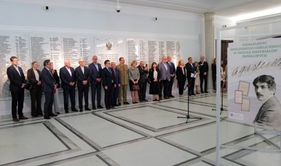 Grupa ludzi w oficjalnych strojach stoi w szeregu wewnątrz jasnej sali z marmurową podłogą. Za nimi znajduje się duża biała ściana z wygrawerowanymi nazwiskami. Na pierwszym planie znajduje się tablica z portretem mężczyzny i polskim tekstem o Apolinarym Grzędzieckim.