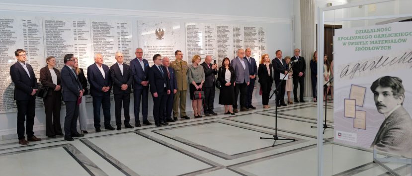 Grupa ludzi w oficjalnych strojach stoi w szeregu wewnątrz jasnej sali z marmurową podłogą. Za nimi znajduje się duża biała ściana z wygrawerowanymi nazwiskami. Na pierwszym planie znajduje się tablica z portretem mężczyzny i polskim tekstem o Apolinarym Grzędzieckim.