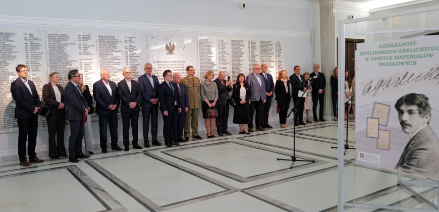 Grupa ludzi w oficjalnych strojach stoi w szeregu wewnątrz jasnej sali z marmurową podłogą. Za nimi znajduje się duża biała ściana z wygrawerowanymi nazwiskami. Na pierwszym planie znajduje się tablica z portretem mężczyzny i polskim tekstem o Apolinarym Grzędzieckim.