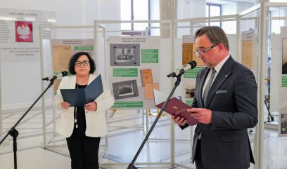 Mężczyzna w garniturze i okularach mówi do mikrofonu, trzymając teczkę. Kobieta w białej marynarce stoi za nim, również trzymając folder i przemawiając. Znajdują się w jasnym pomieszczeniu z tablicami informacyjnymi w tle.