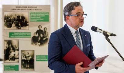 Mężczyzna w niebieskim garniturze i okularach mówi do mikrofonu, trzymając czerwoną teczkę. Za nim znajduje się tablica z czarno-białymi zdjęciami i polskim tekstem, ustawiona w jasnym, nowoczesnym wnętrzu z białymi ścianami.