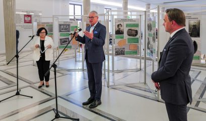 Trzy osoby w formalnych strojach stoją w jasnej przestrzeni wystawowej z historycznymi tablicami; jeden mężczyzna mówi do mikrofonu na podium, podczas gdy drugi mężczyzna i kobieta stoją obok i słuchają. Pokój ma marmurowe podłogi i białe ściany.