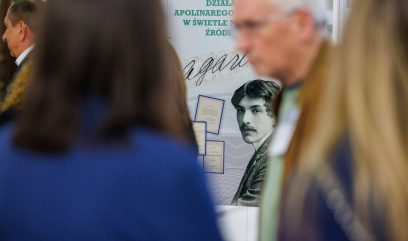 Niewyraźny tłum stoi przed wyświetlaczem przedstawiającym czarno-biały portret wąsatego mężczyzny o ciemnych włosach. Polski tekst i dokumenty historyczne są widoczne na plakacie za nimi. W centrum uwagi jest wyświetlacz, a nie ludzie.