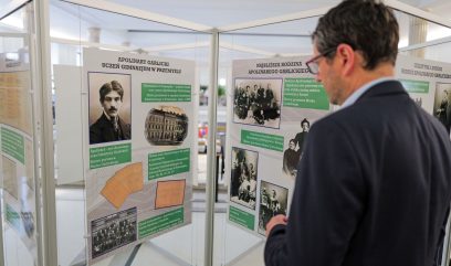 Mężczyzna w garniturze z krótkimi ciemnymi włosami i w okularach czyta tablice muzealne zawierające historyczne zdjęcia, dokumenty i polski tekst o Apolinarym Gałeckim. Tablice są zamontowane na białych ramach w jasno oświetlonym wnętrzu.