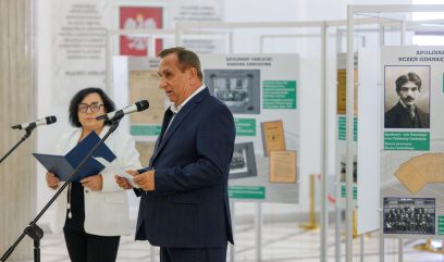 Mężczyzna w niebieskim garniturze przemawia do mikrofonu, trzymając dokumenty. Kobieta w białej marynarce i czarnych spodniach stoi obok niego, również trzymając dokumenty. Znajdują się w przestrzeni wystawowej z tablicami informacyjnymi w tle.