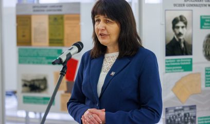 Kobieta o prostych ciemnych włosach w niebieskiej marynarce przemawia do mikrofonu. Za nią znajdują się tablice informacyjne z tekstem, historycznymi zdjęciami i dokumentami. Sceneria wydaje się być wystawą lub formalnym wydarzeniem w dobrze oświetlonym pomieszczeniu.