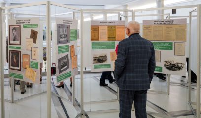 Starszy mężczyzna w garniturze w kratę stoi przed tablicami informacyjnymi ze zdjęciami i dokumentami na wystawie wewnątrz budynku. Inni ludzie oglądają pobliskie ekspozycje. Wystawa znajduje się w jasnym, przestronnym pomieszczeniu z białymi ścianami i czarnymi liniami podłogi.