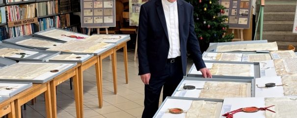 Starszy mężczyzna o białych włosach, w okularach i ciemnym garniturze, stoi w bibliotece i przygląda się historycznym dokumentom rozłożonym na stołach. Za nim znajdują się półki z książkami, plakaty informacyjne i udekorowana choinka.