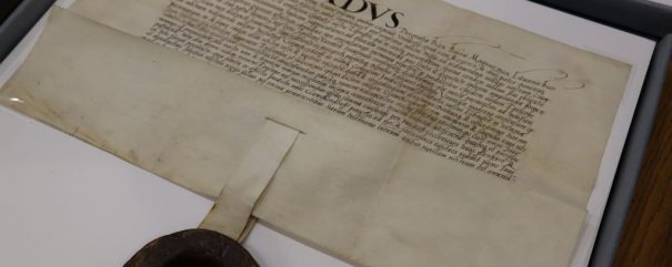 Historyczny dokument zatytułowany SIGISMUNDUS, napisany ozdobnym pismem łacińskim na postarzanym pergaminie. Duża czerwona pieczęć woskowa jest przymocowana szerokim paskiem pergaminu, a dokument jest wyświetlany na białym tle.