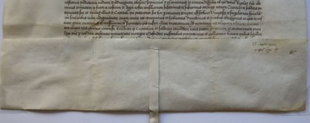 Średniowieczny dokument pergaminowy z kaligraficznym pismem łacińskim, z ozdobnymi czarnymi literami u góry. Dolna krawędź ma wstążkę z czerwoną pieczęcią woskową, prawdopodobnie oznaczającą autentyczność lub autorytet. Pergamin wykazuje pewne zagięcia i niewielkie przebarwienia.