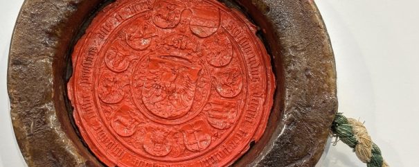 Duża, okrągła średniowieczna czerwona pieczęć woskowa ze skomplikowanymi wzorami i inskrypcjami, zamknięta w grubym brązowym pierścieniu ochronnym. Do pieczęci przymocowany jest zielono-biały pleciony sznur, spoczywający na jasnej powierzchni.