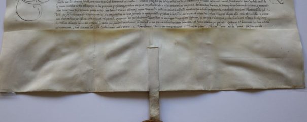 Historyczny dokument pergaminowy z ozdobnym pismem łacińskim i wyszukaną kaligrafią u góry. Woskowa pieczęć w okrągłym etui zwisa ze wstążki przymocowanej do dolnej części pergaminu. Papier jest postarzony i lekko pożółkły.