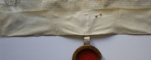 Historyczny dokument pergaminowy z ozdobnym, kaligraficznym pismem u góry, zaczynającym się od STEPHANV. Poniżej znajduje się duża czerwona pieczęć woskowa w okrągłym uchwycie, przymocowana dwiema kremowymi wstążkami. Pergamin wykazuje pewne zagniecenia i starzenie się.