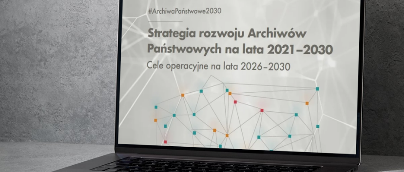 Laptop ustawiony na szarym blacie przed betonową ścianą, na ekranie dokument w języku polskim z nagłówkiem „Strategia rozwoju Archiwów Państwowych na lata 2021–2030”, podtytułem „Cele operacyjne na lata 2026–2030”, widocznym hashtagiem „#ArchiwaPaństwowe2030” oraz schematyczną grafiką sieci kolorowych punktów połączonych liniami.