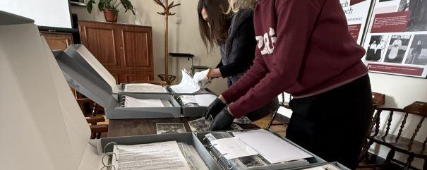 Kilka szarych pudeł archiwalnych z segregatorami i czarno-białymi zdjęciami rozłożonych na stole, przy którym dwie kobiety — jedna w bordowej bluzie i czarnych rękawiczkach pochylona nad fotografiami, druga w ciemnym swetrze przewracająca kartki — przeglądają materiały w pomieszczeniu z drewnianą szafką i stojącym wieszakiem, rośliną doniczkową, kinkietem na ścianie oraz planszami wystawowymi z historycznymi zdjęciami.