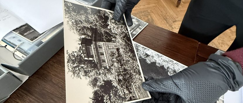Dłonie w czarnych rękawiczkach trzymają czarno-białe odbitki przedstawiające budynek wśród drzew, ułożone na ciemnym drewnianym stole obok otwartego szarego pudełka z przezroczystymi koszulkami na zdjęcia, w pomieszczeniu z parkietem i widoczną nogą krzesła.
