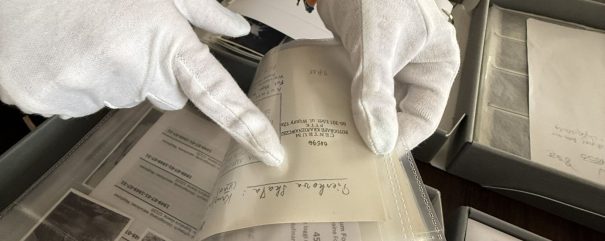 Dwie dłonie w białych bawełnianych rękawiczkach, jedna z pomarańczową opaską na nadgarstku, trzymają i wskazują przezroczystą foliową koszulkę z dokumentami i kartami z odręcznymi zapisami, a wokół leżą pudełka z czarno-białymi fotografiami i kopertami.
