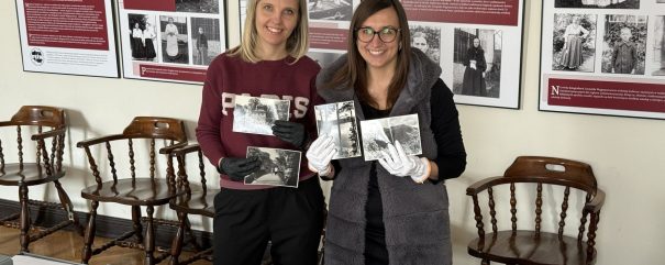 Dwie kobiety stoją na tle plansz wystawy ze starymi czarno‑białymi fotografiami i opisami, między drewnianymi krzesłami, trzymają w rękach historyczne czarno‑białe zdjęcia — kobieta po lewej w bordowej bluzie i czarnych rękawiczkach, kobieta po prawej w okularach, ciemnej kamizelce i białych rękawiczkach, na pierwszym planie stół z otwartymi pudełkami i dokumentami.