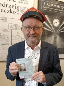 Mężczyzna z siwą brodą i okularami w czerwonej czapce i granatowej marynarce trzyma oboma rękami legitymację ze zdjęciem, a w tle widoczne plakaty z napisami „Stacja Muzeum” i czarno-białym zdjęciem peronu z zegarem.