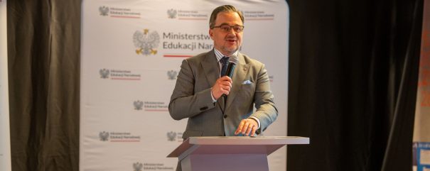 Mężczyzna w okularach i szarym garniturze stoi za pulpitem i trzyma mikrofon na tle białego baneru z godłem i napisem „Ministerstwo Edukacji Narodowej” oraz ciemnych zasłon.