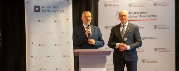 Dwóch mężczyzn w garniturach stoi za mównicą na scenie trzymając mikrofony, za nimi banery z napisami „Archiwa Państwowe” i „Ministerstwo Edukacji Narodowej”, a na przedzie mównicy widoczny baner z napisem „Bez korzeni nie zakwitniesz”.