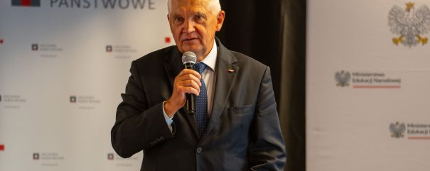 Starszy mężczyzna z siwymi włosami w ciemnym garniturze i niebieskim krawacie, stojący za mównicą i trzymający mikrofon, na tle banerów z napisem „Archiwa Państwowe” oraz fragmentami godła i logo Ministerstwa Edukacji Narodowej.