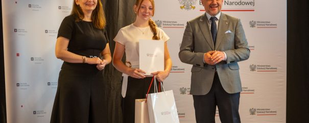 Trzy osoby stoją przed banerami z napisami „Archiwa Państwowe” i „Ministerstwo Edukacji Narodowej”: kobieta po lewej w czarnej sukience, młoda kobieta pośrodku trzymająca dokument i białe torby upominkowe oraz mężczyzna po prawej w szarym garniturze i granatowym krawacie.