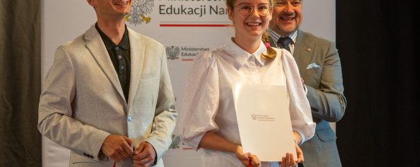 Dwóch mężczyzn i młoda kobieta stoją przed banerem z napisem Ministerstwo Edukacji Narodowej i godłem; kobieta trzyma biały folder z logo ministerstwa, na pierwszym planie widoczne białe torby z czerwonymi uchwytami.